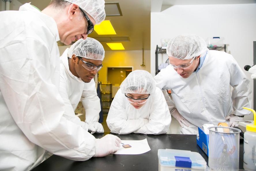 Four people wearing lab equipment standing around a table, looking at a microchip