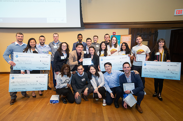 Winners from the $100k New Ventures Competition pose for a group photo with their large prop checks