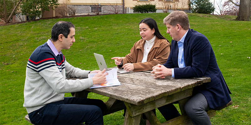 Bridget Moynihan, E21, sits at a picnic table and looks at a computer screen with Professor of the Practice Eric Hines while Professor Babak Moaveni sits on the other side of the picnic table looking at a book.