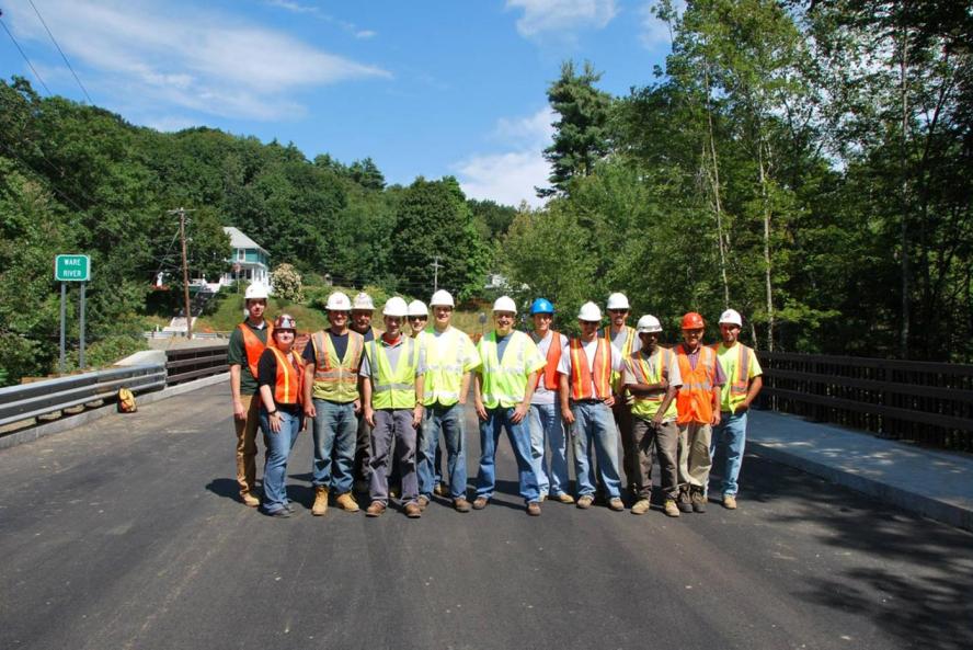 Men and women wearing safety vests and safety helmets, standing on roadway