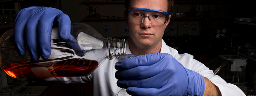 Scientist, wearing protective goggles and rubber gloves, pouring liquid from a bottle into a smaller vessel.