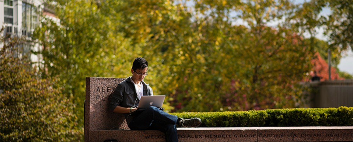 Student, sitting outdoors on concrete bench, looking at laptop.
