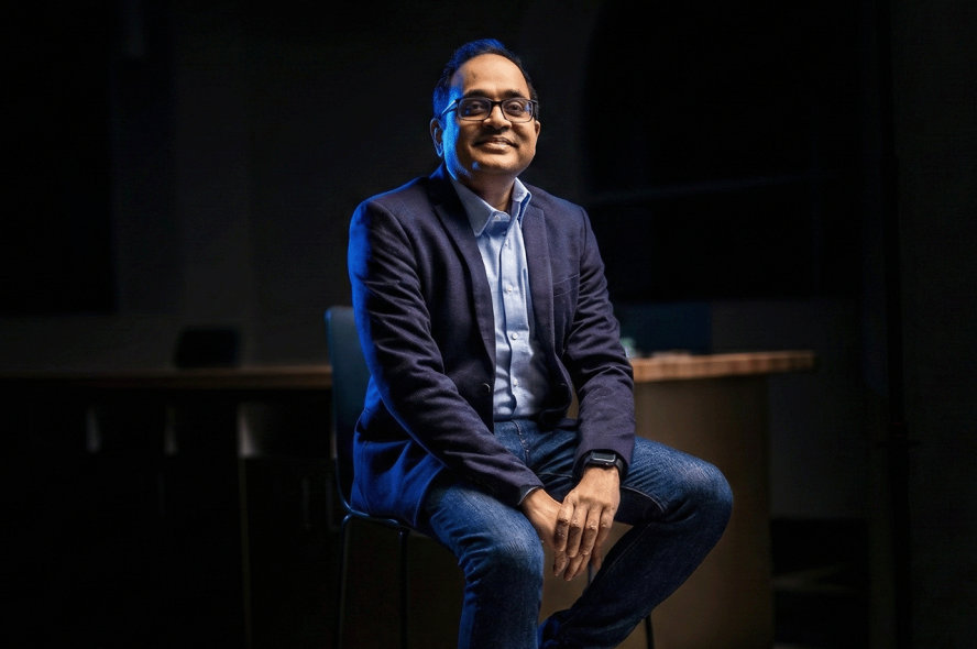 A confident man wearing glasses and a blue suit jacket over a light blue shirt and jeans is seated on a high stool in a dimly lit, contemporary office space with blue lighting accents.