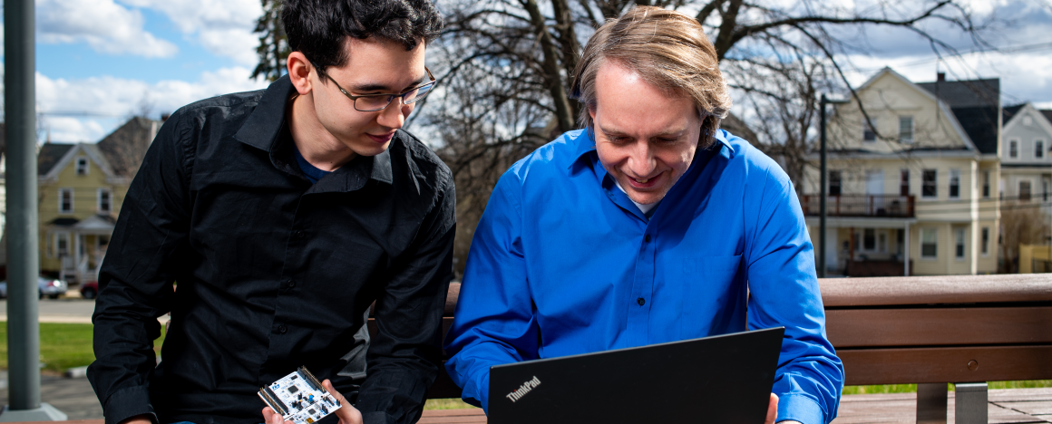 Associate Professor Robert White and mechanical engineering student Tim Cheng look at a laptop together. Tim is holding a piece of mechanical engineering equipment. 