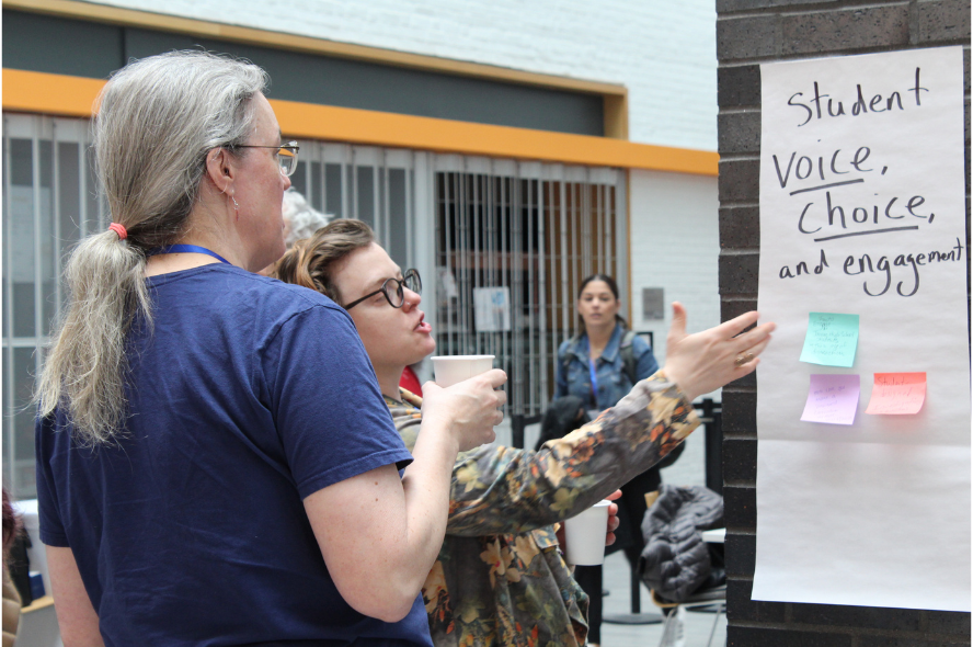 Two attendees at the Eye on STEAM conference look and point at a poster that reads "Student Voice, Choice and Engagement" and gesture towards some colorful sticky notes on the poster.
