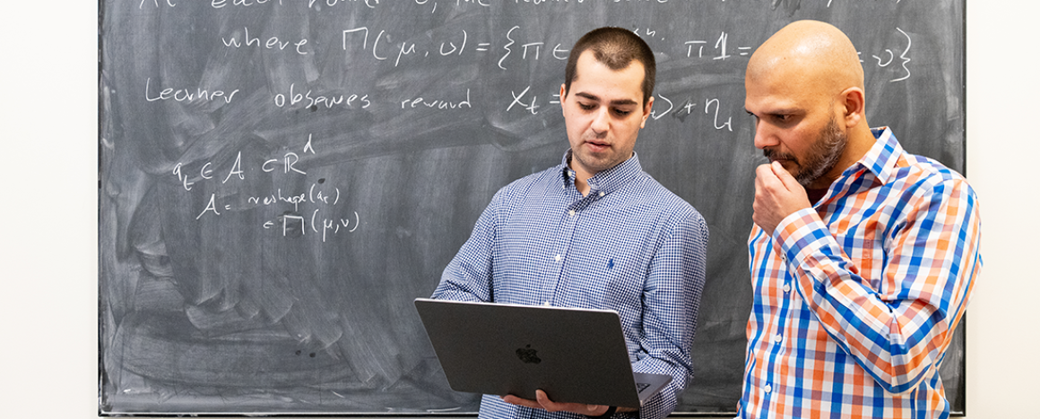 Sandro Avaliani (left) and Shuchin Aeron (right) look at a laptop together. They are standing in front of a blackboard with many equations written on it.