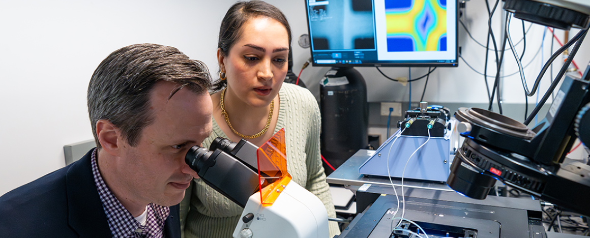 Jeff Guasto (left) looks in a microscope as Maliheh Teimouri looks on.