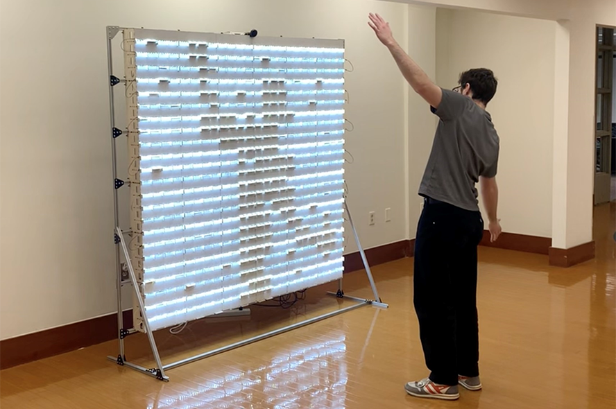Student standing in front of art exhibition with arms outstretched. The same shadow shows up in the art piece.