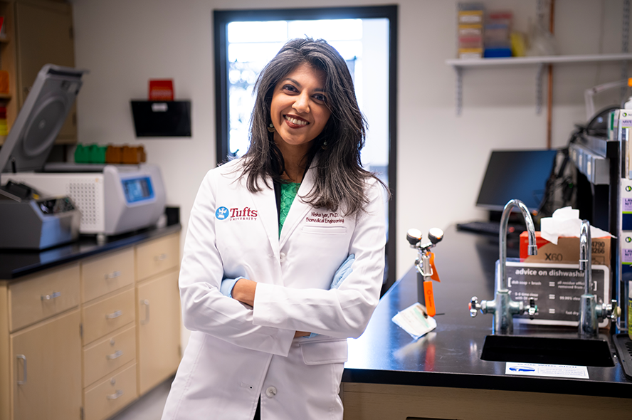 Assistant Professor Nisha Iyer in her lab wearing a white lab coat with a Tufts University logo. 