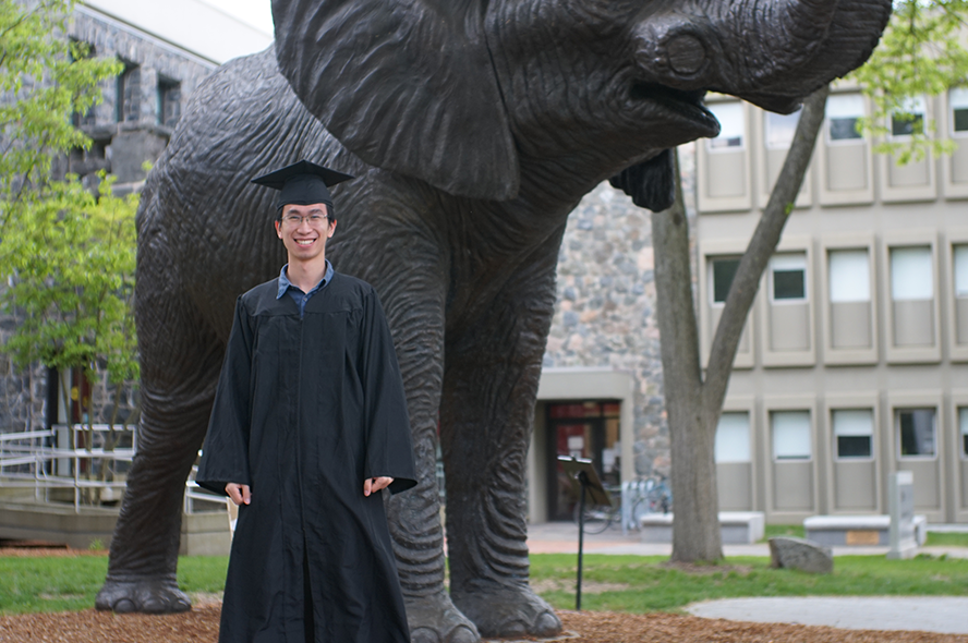 Tufts alum Eric Wu wearing graduation robes and a graduation cap. He is standing in front of the Jumbo statue on the Tufts Medford campus. 