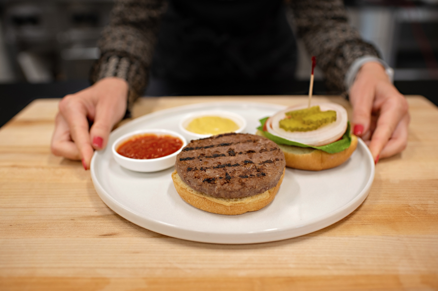 An open-faced hamburger on a bun, on a white plate along with a burger top with fixings, atop a wood table, with a woman’s arms on either sides of the plate.