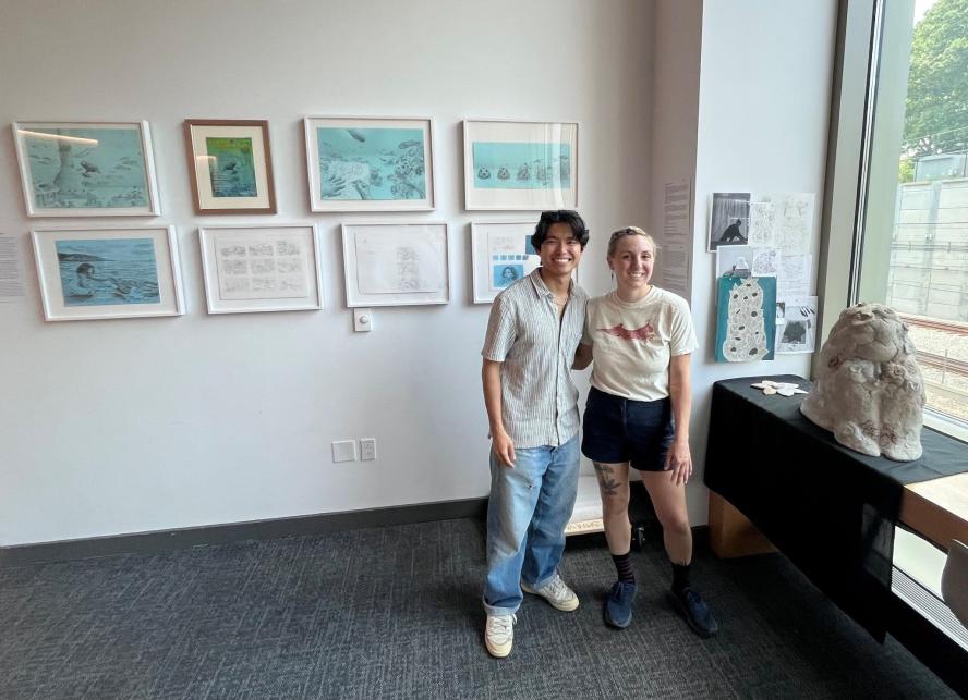 A young man and women stand in front of a small gallery of artwork
