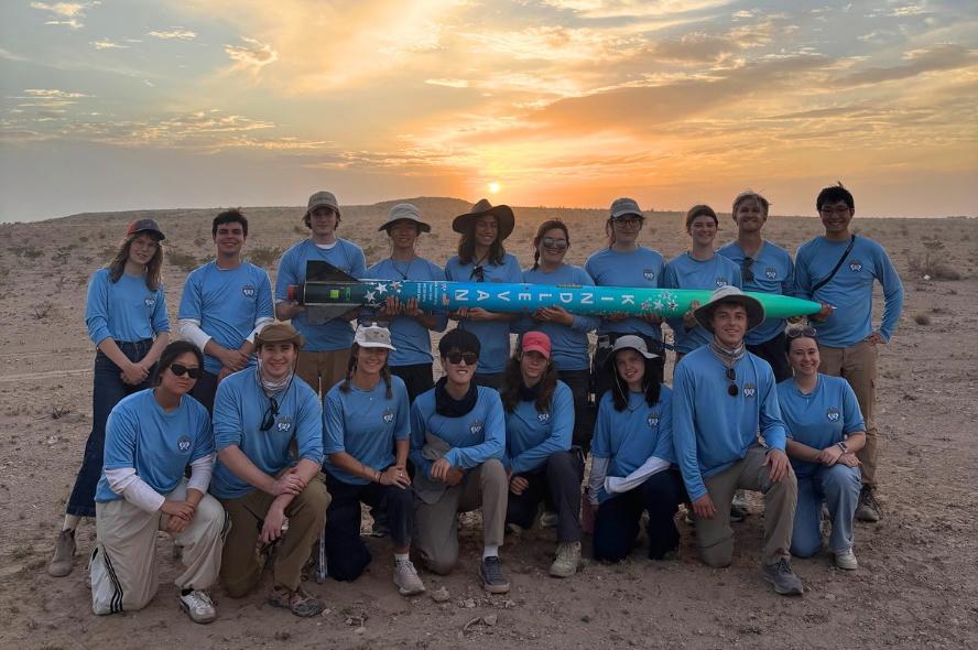 Large group of young adults holding blue rocket in a desert environment