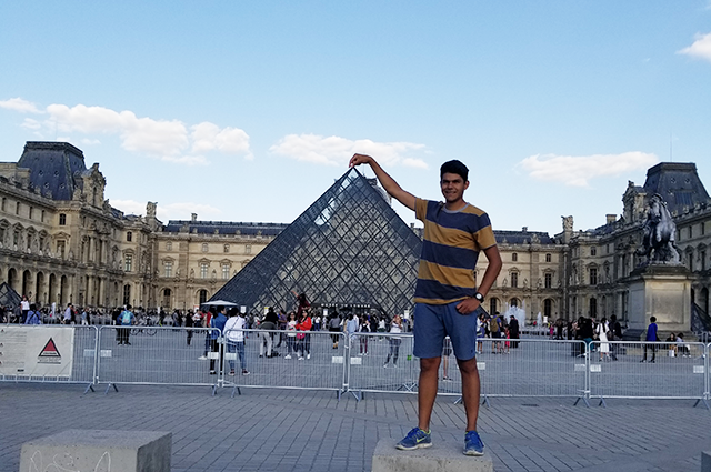 A college student in shorts and a T-Shirt pretends to pinch the top of the Louvre glass triangle.
