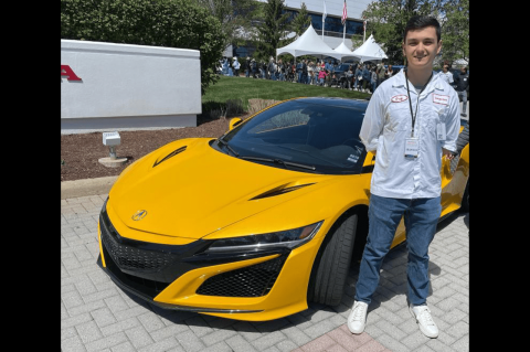 A young man wearing a lanyard stands next to a bright yellow car.