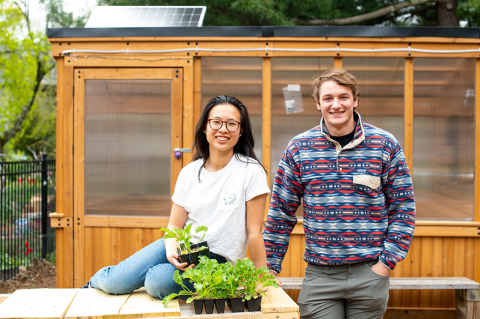 Two students with plants posing and smiling in front of solar-powered greenhouse