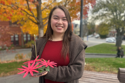 Madison Giles stands outside against a background of fall foliage. She is holding her hummingbird feeder which mimics the red passionflower.