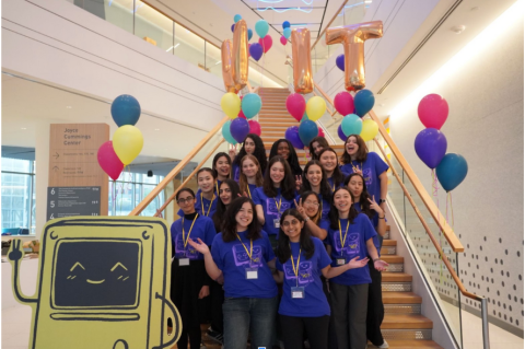 A group of roughly 20 Women in Tech conference organizers and attendees pose for a photo on the Joyce Cummings Center steps. They are all wearing purple t-shirts. Behind them are many balloons including three gold balloons that read "WIT."