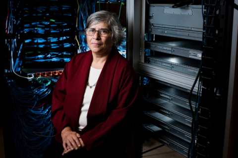 A white woman with short grey hair sits in a dark room next to a computer.