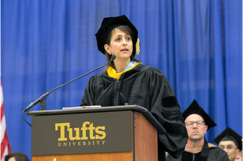 A white woman with brown hair stands at a Tufts podium in academic regalia