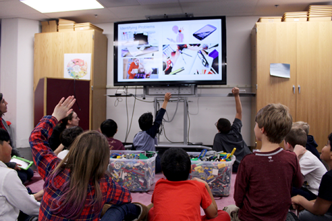 Elementary school children looking at a television screen, displaying information that they submitted to the Design Keeper app. The children are sitting on the floor and raising their hands.