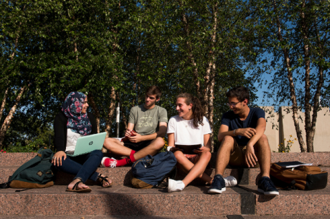 Four smiling students with laptops sit together outside on a sunny day