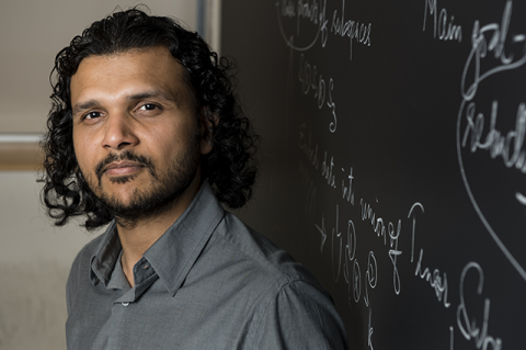 A brown man with shoulder-length curly black hair stands against a chalkboard with writing on it.