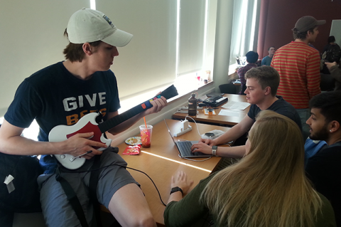 Student seated on a table plays a toy guitar hooked up to a laptop, as other students work with the laptop and look on